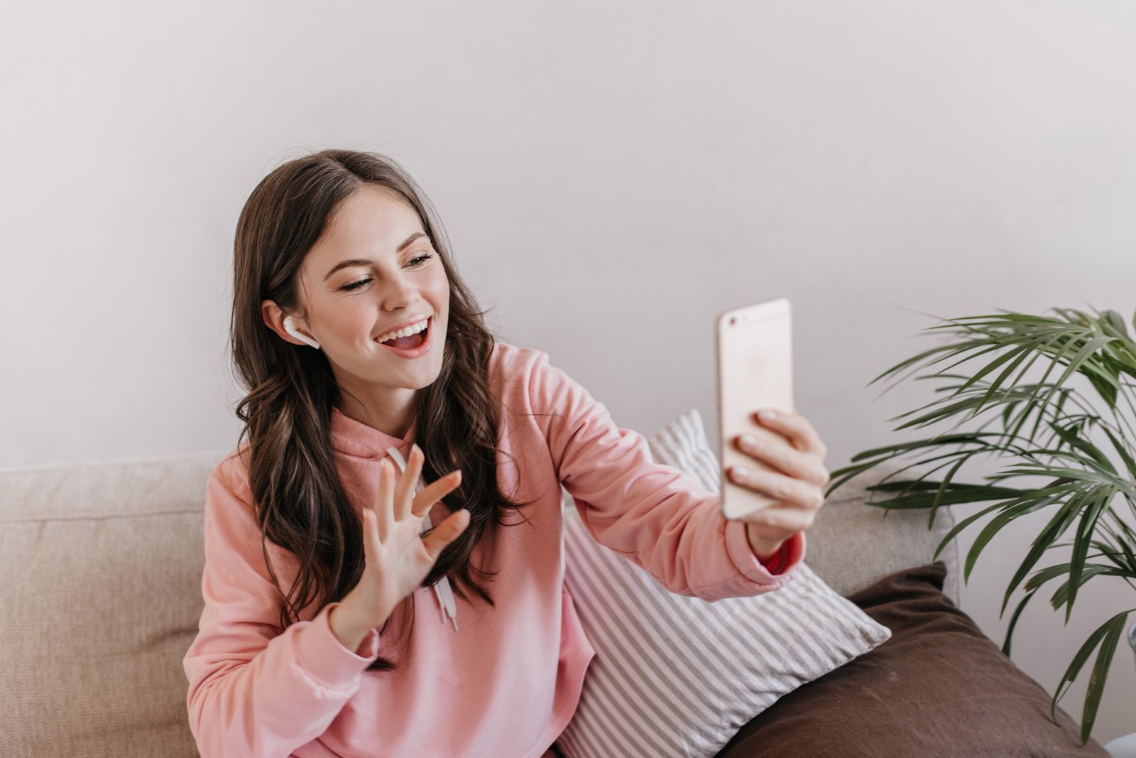 Positive girl in pink sports outfit talking on phone in wireless headphones and sitting on couch. Happy woman in pink hoodie smiles and has video call with friend